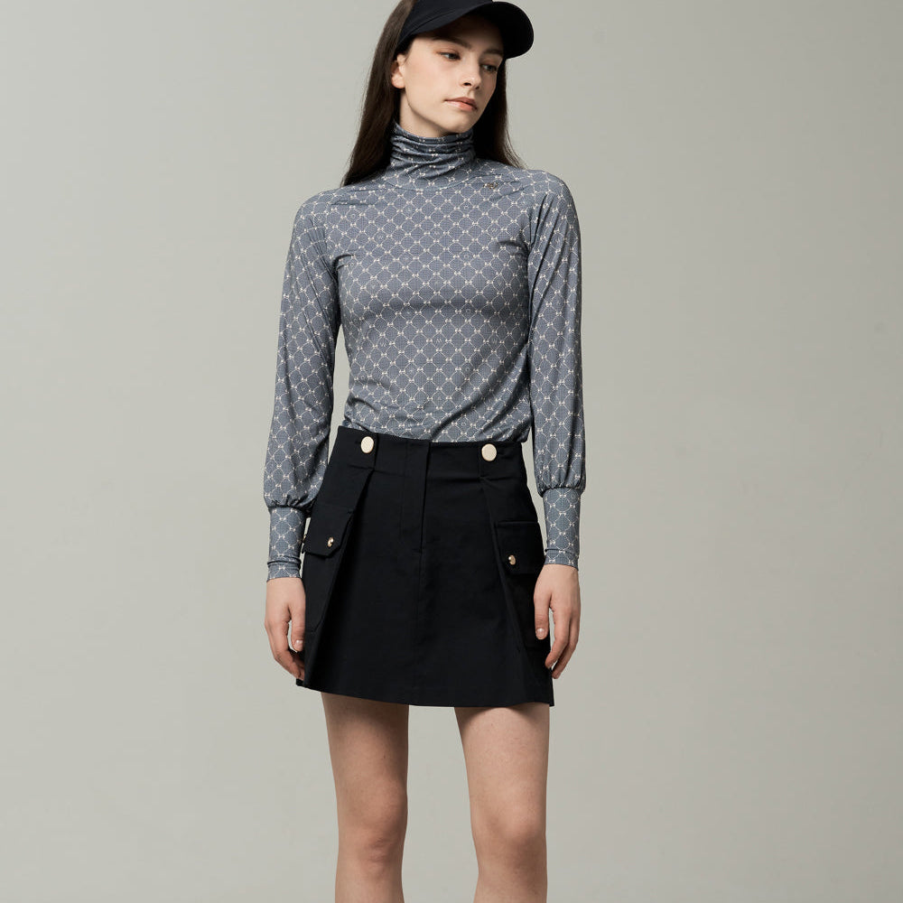A model stands against a light gray studio background wearing a gray patterned long-sleeve mock-neck top, a black pleated mini skirt with gold-tone buttons, a black visor, white socks, and black-and-white sneakers.