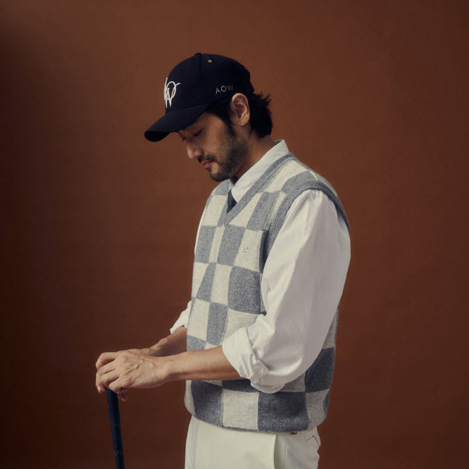 A man in a black baseball cap, a gray and white checkered knit sweater vest over a white shirt, and white trousers holding a golf club against a solid dark brown background.
