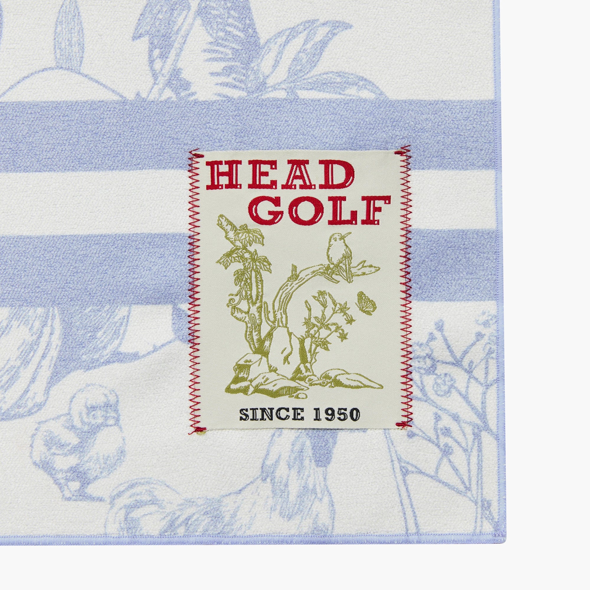 Close-up of a square golf towel or mat with a white base, light blue botanical stripes, and a central cream patch reading 'HEAD GOLF' with red stitching and green illustration. The product fills most of the frame against a white background.