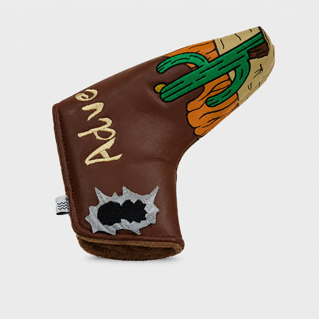 A brown leather golf club headcover featuring a desert-themed design with a green cactus, orange sun, and a silver bullet hole patch, on a light gray background.