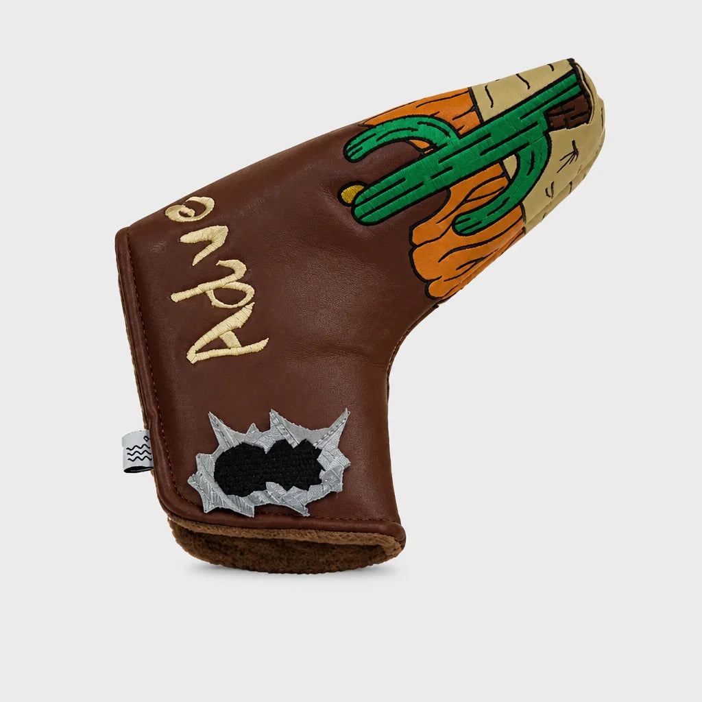 A brown leather golf club headcover featuring a desert-themed design with a green cactus, orange sun, and a silver bullet hole patch, on a light gray background.