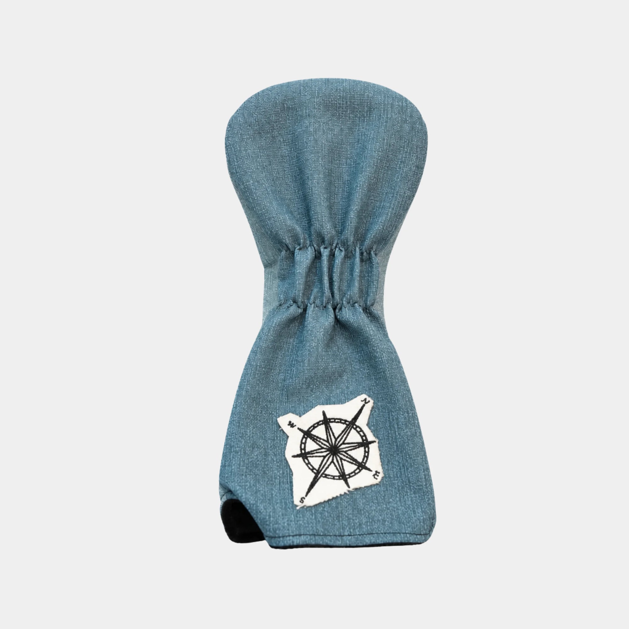 A blue denim-textured golf club headcover with a white compass rose patch on the lower front and a black rubber thumb hole, shown against a light gray background.