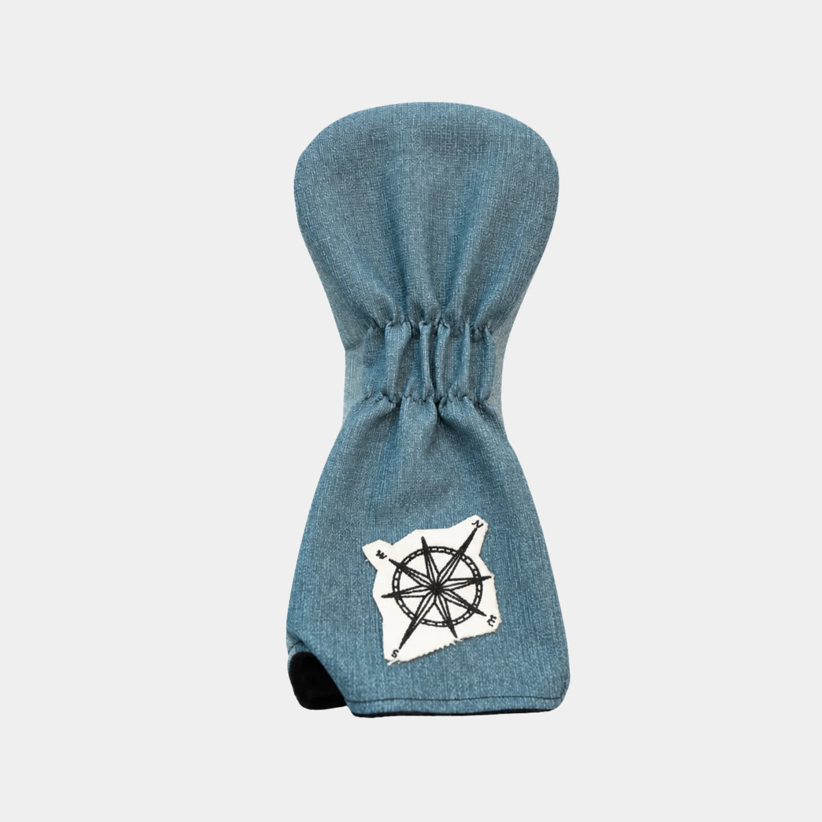 A blue denim-textured golf club headcover with a white compass rose patch on the lower front and a black rubber thumb hole, shown against a light gray background.