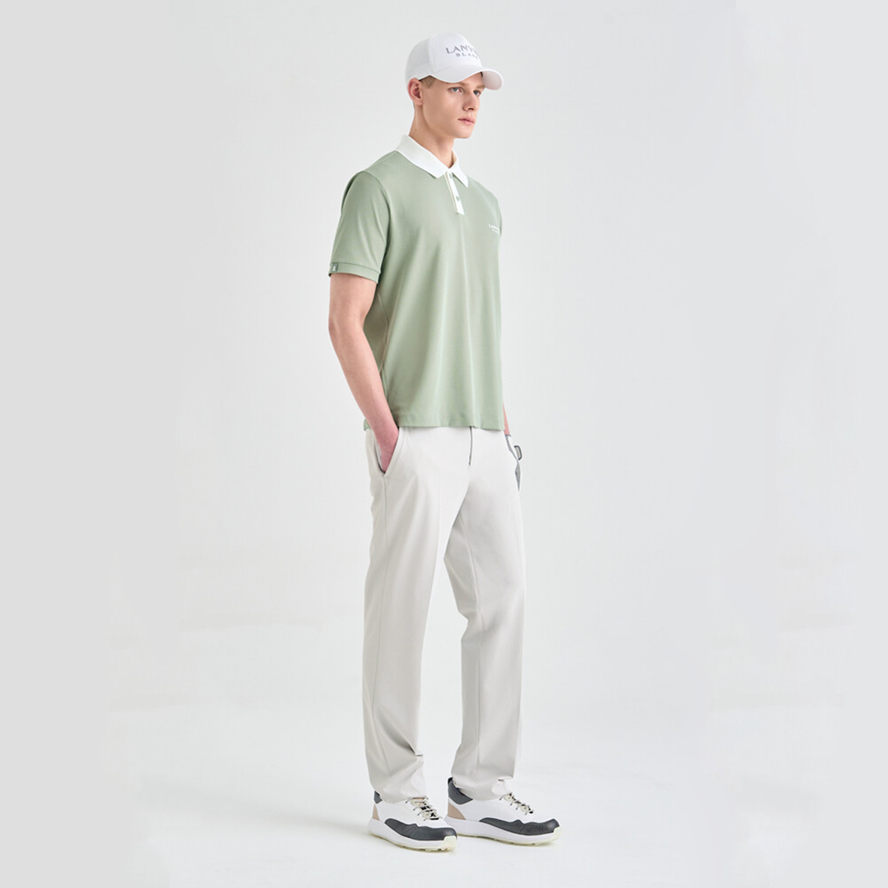 Man wearing a light green polo shirt, white pants, and a white cap on a plain background