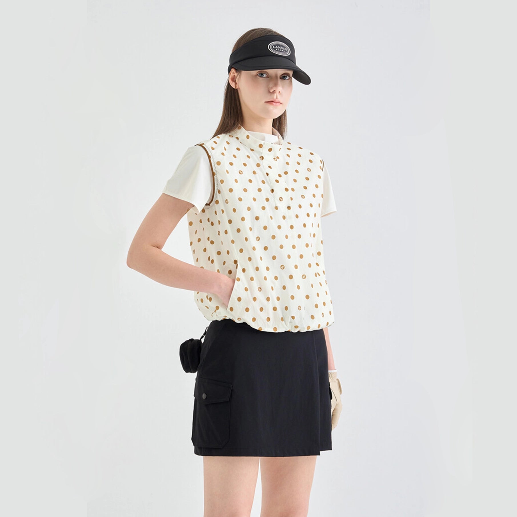 Woman wearing a white blouse with gold polka dots and black cap on a light gray background