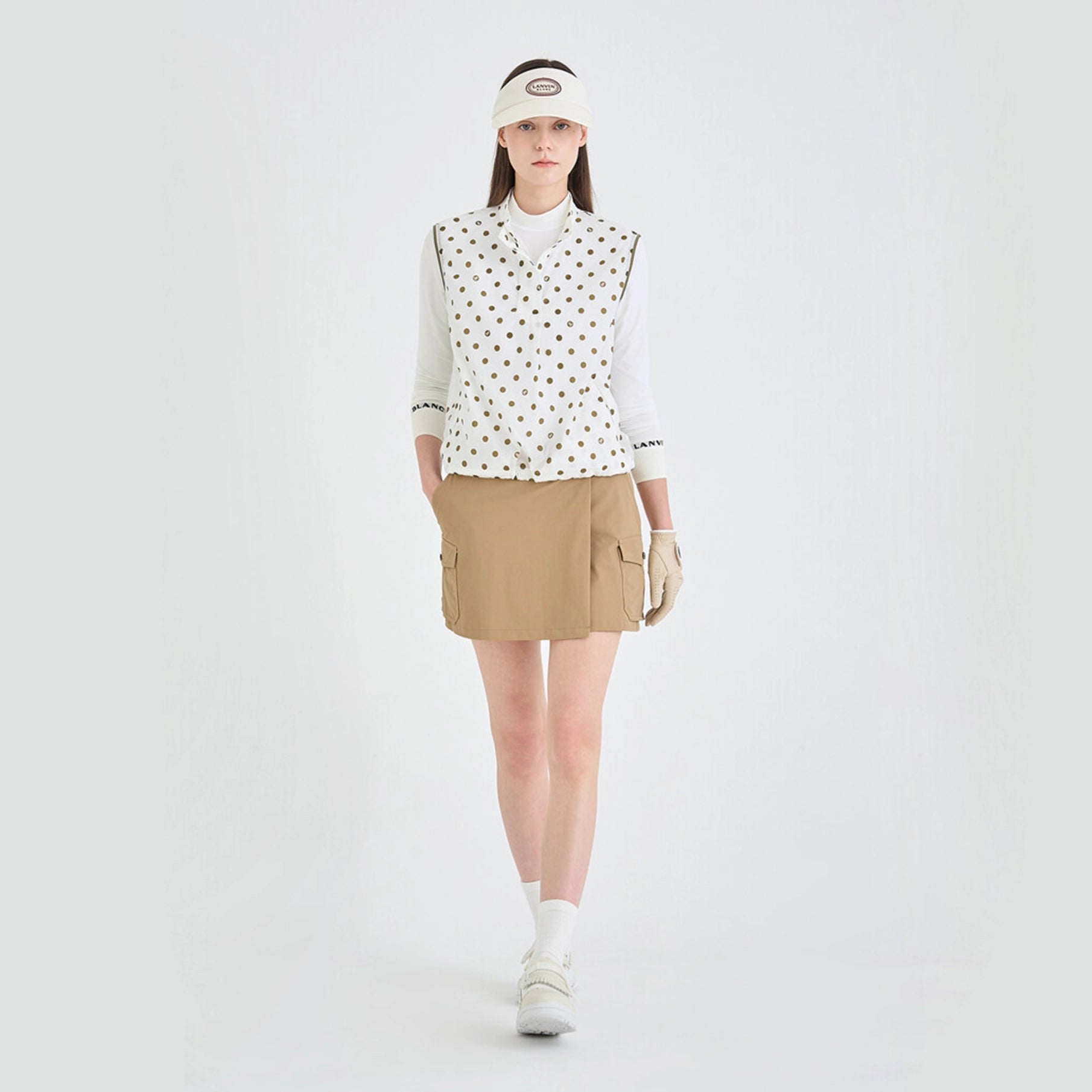 Woman wearing a polka dot shirt, beige skirt, and white visor on a white background