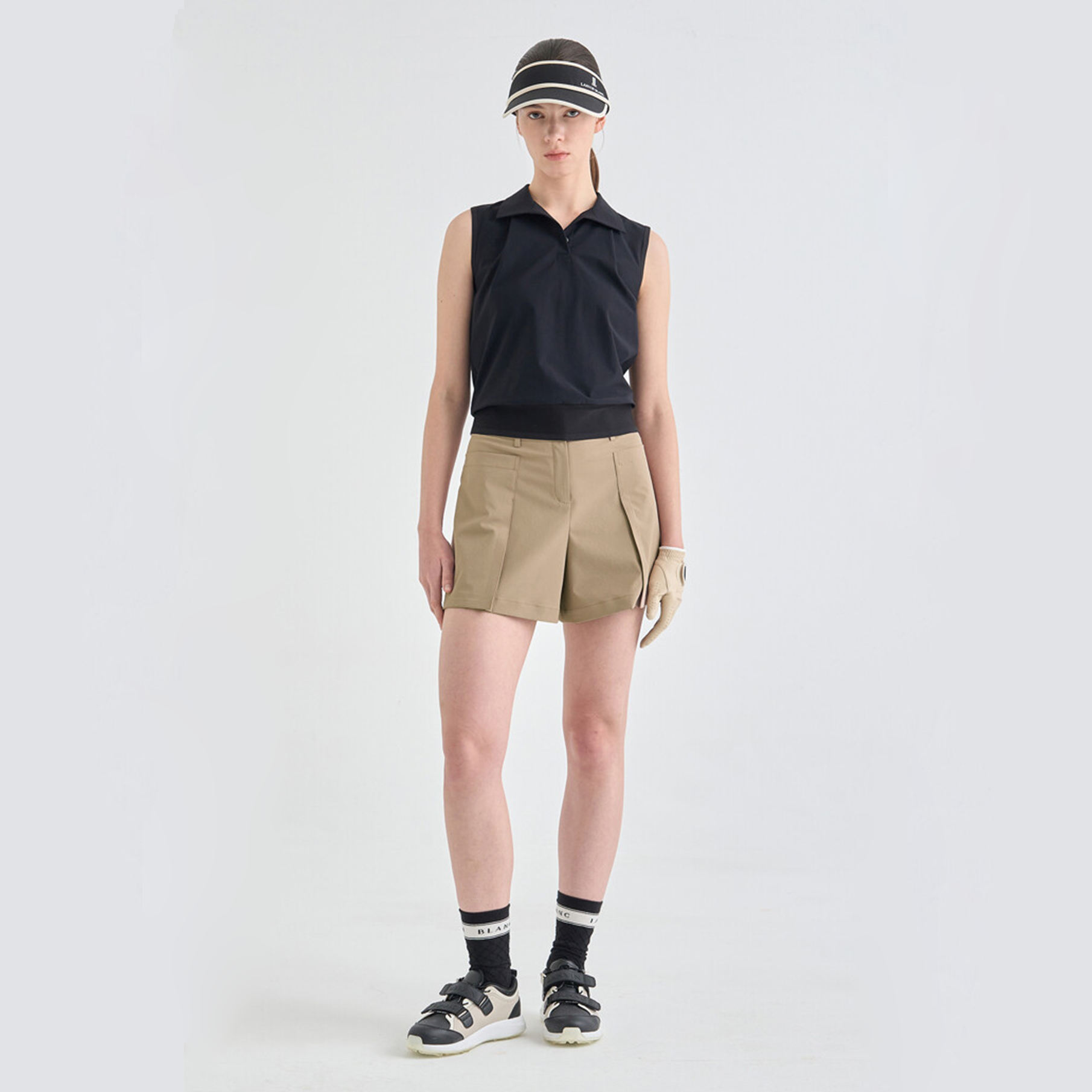 Person wearing a black sleeveless top, beige shorts, and black shoes on a white background