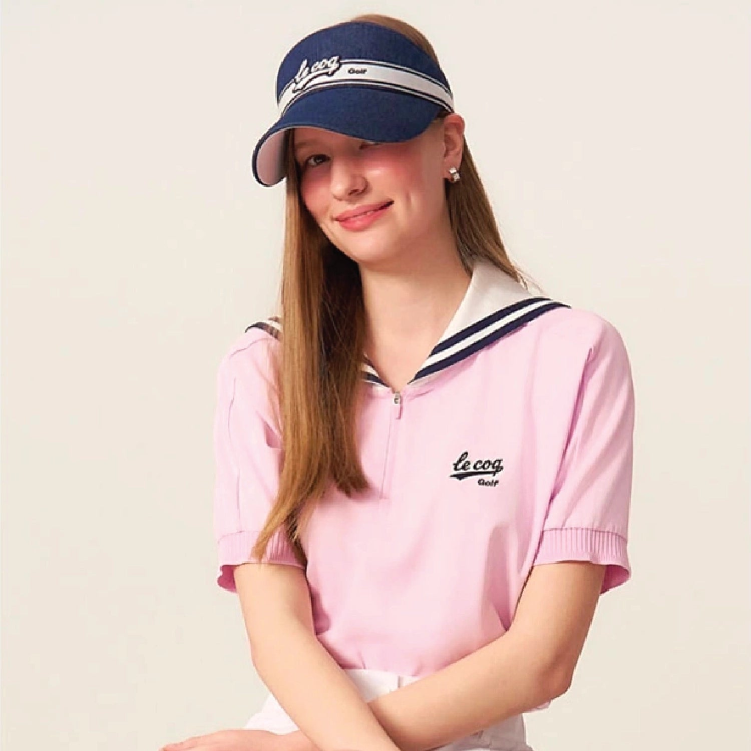 A young woman wearing a pink short-sleeve golf polo shirt with a white collar and navy trim, paired with a navy blue visor. The brand name 'le coq Golf' is embroidered on the chest and visor.