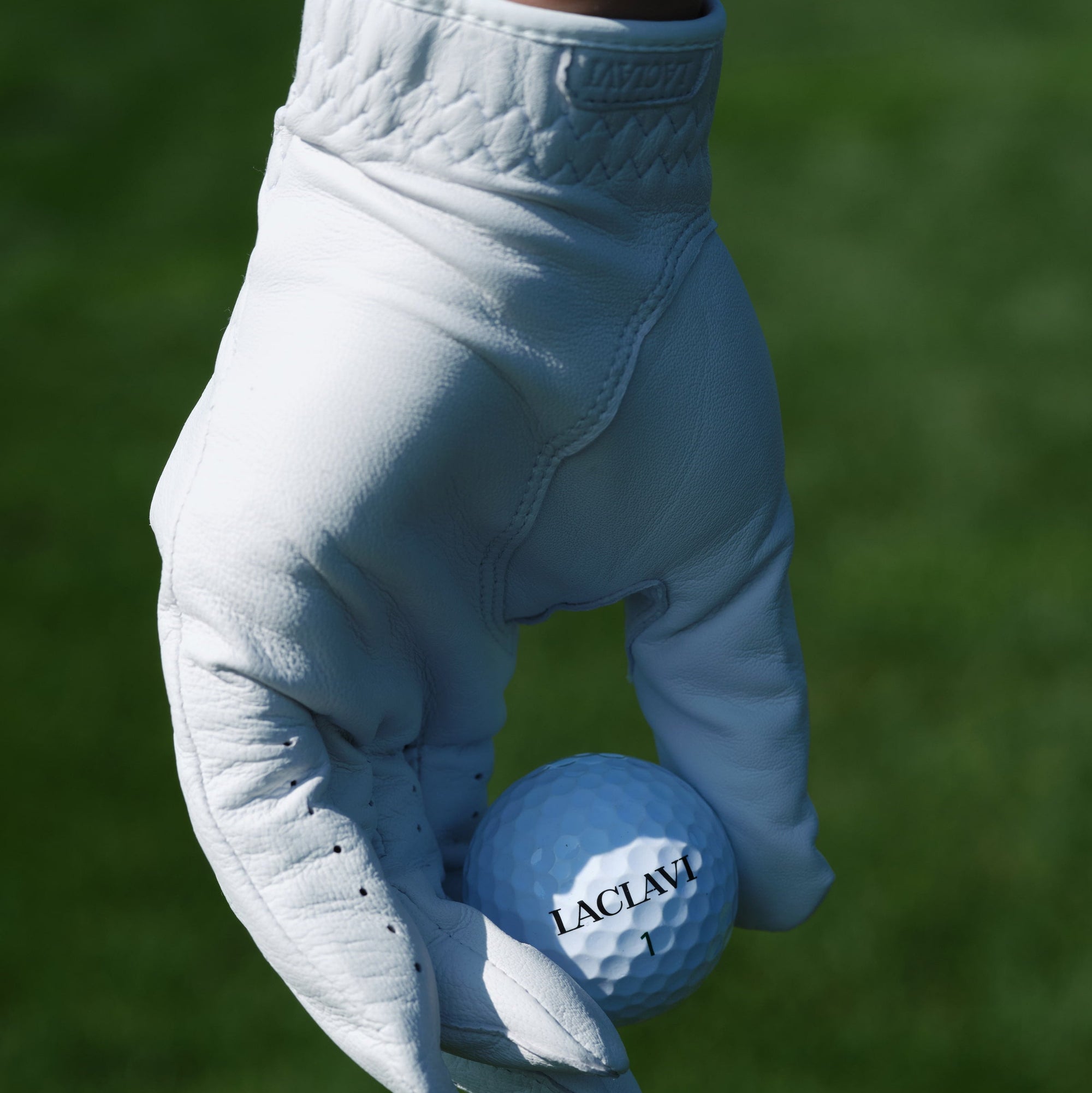 Golfer's hand in white cabretta tour golf glove holding Laclavi branded golf ball on green grass - Laclavi Golf