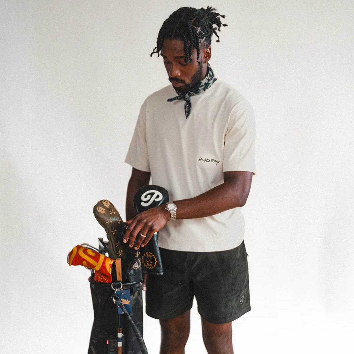 A man wearing a cream T-shirt standing with his golf clubs.