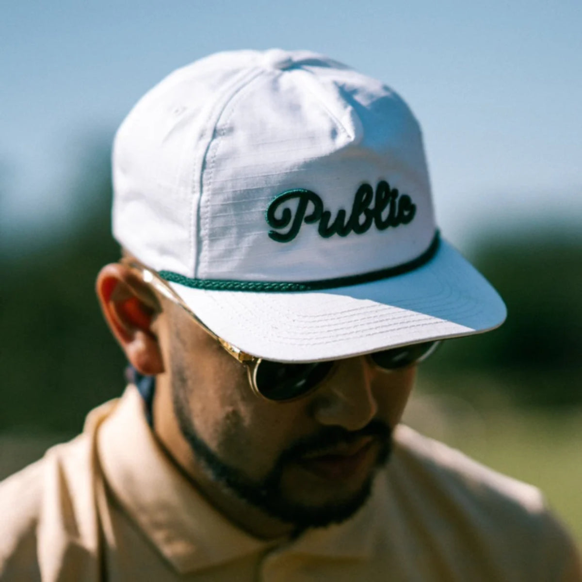 A man wearing a white cap with 'Public' branding and sunglasses, outdoors.
